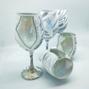 May include: Four wine glasses with a zebra print design and iridescent glitter accents. The glasses are arranged in a group on a white surface.