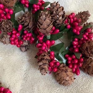 May include: Close-up of two small, decorative wreaths. Each wreath features brown pinecones, bright pink berries, and green leaves. The wreaths are arranged on a neutral-colored, textured surface, suggesting a seasonal or holiday theme.