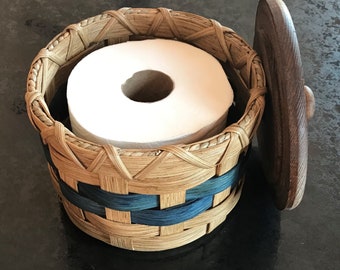 May include: A woven basket with a blue and brown pattern holds a roll of white toilet paper. The basket has a separate lid made of dark wood.