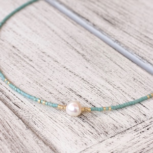 May include: A delicate necklace featuring a single white freshwater pearl centred on a strand of small, matte teal seed beads. The design is accented with tiny, metallic gold-toned cylindrical spacer beads interspersed along the teal bead strand.