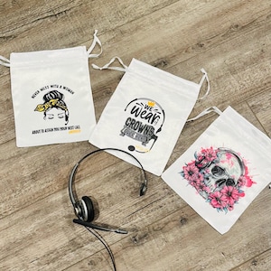 Peut inclure: Trois sacs blancs à cordon avec des motifs graphiques et un casque. Un sac porte l'inscription "NEVER MESS WITH A WOMAN" avec un visage de femme. Un autre dit "WE WEAR CROWNS OVER HERE". Le troisième sac a un crâne avec des fleurs roses.