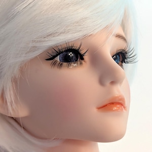 May include: A doll head featuring smooth, pale skin, bright blue eyes with long, dark, defined eyelashes, and short, straight white hair. The facial features include a small, soft nose and glossy, peach-colored lips, captured in a profile view.