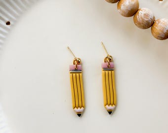 May include: A pair of yellow pencil-shaped earrings with pink erasers and black tips. The earrings have gold-colored ear wires and small gold beads. The earrings are on a white plate with a wooden bead garland.