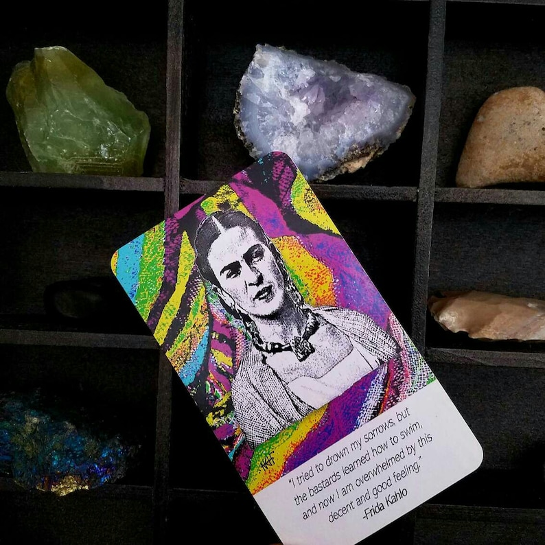May include: A colorful card featuring a portrait of Frida Kahlo with a quote. The card has a vibrant rainbow background. The quote reads, "I tried to drown my sorrows, but the bastards learned how to swim, and now I am overwhelmed by this decent and good feeling."