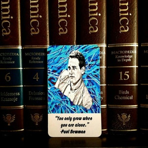 May include: A rectangular card featuring a portrait of Paul Newman against a blue abstract background. The card includes the quote, "You only grow when you are alone." The card is set against a backdrop of brown encyclopedias.