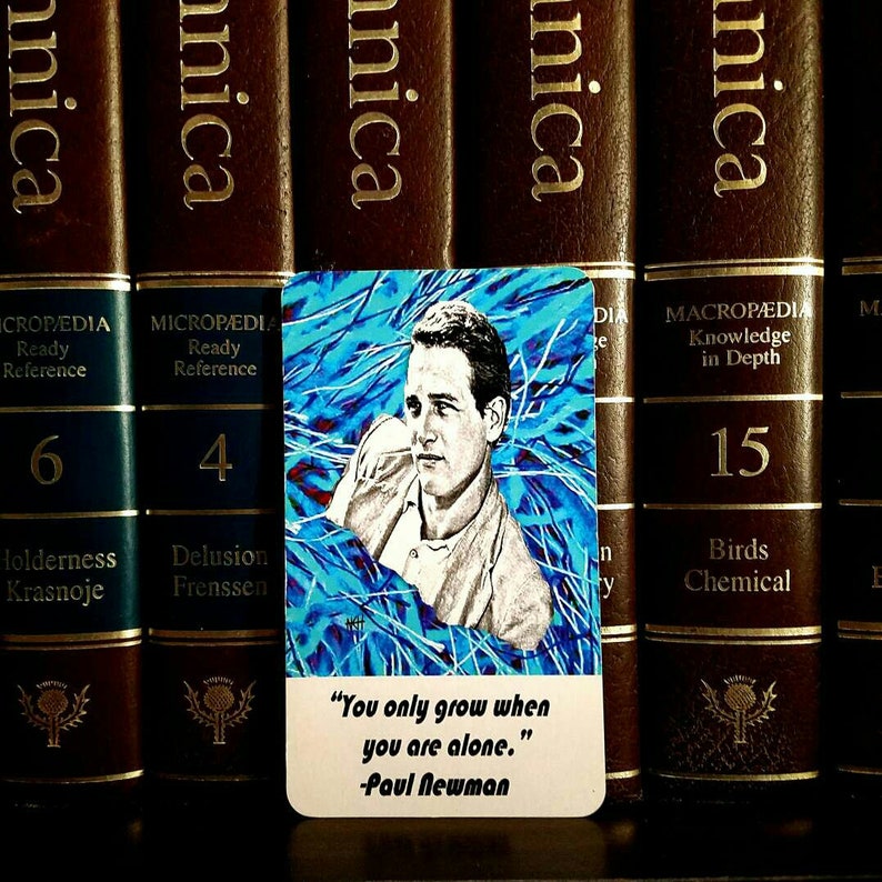 May include: A rectangular card featuring a portrait of Paul Newman against a blue abstract background. The card includes the quote, "You only grow when you are alone." The card is set against a backdrop of brown encyclopedias.
