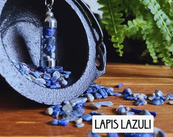 May include: A silver-toned cylindrical pendant filled with small, tumbled blue and white lapis lazuli stones, hanging from a chain. The pendant is positioned inside a black cast iron cauldron, with loose lapis lazuli stones scattered nearby. The text LAPIS LAZULI is visible.