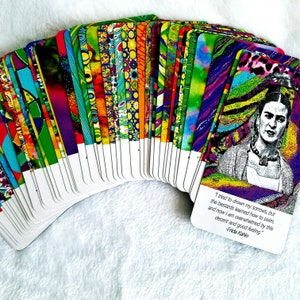 May include: A deck of colorful tarot cards fanned out on a white surface. The card backs feature a variety of vibrant patterns. One card displays a portrait of Frida Kahlo with a quote: "I tried to drown my sorrows, but the bastards learned how to swim..."
