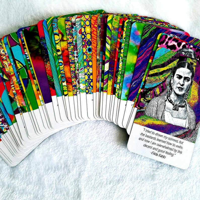 May include: A deck of colorful tarot cards fanned out on a white surface. The card backs feature a variety of vibrant patterns. One card displays a portrait of Frida Kahlo with a quote: "I tried to drown my sorrows, but the bastards learned how to swim..."