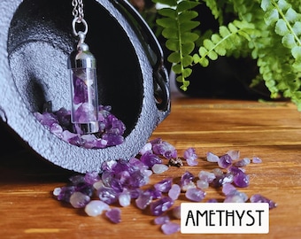 May include: A silver-toned pendant necklace featuring a clear glass vial filled with small, raw purple amethyst stones. The pendant hangs from a black cast iron cauldron, with additional loose amethyst stones scattered nearby and the text AMETHYST.