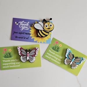 Puede incluir: Tres imanes decorativos: una abeja y dos mariposas, cada uno sobre una tarjeta pequeña. La abeja es amarilla y negra, las mariposas son grises, rosas y azules. Las tarjetas tienen el texto: "¡Gracias por apoyar a nuestra pequeña empresa!"