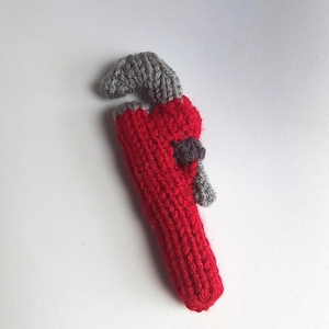 May include: A handmade, knitted adjustable wrench in red and gray. The wrench features a red handle and a gray adjustable jaw. A small, dark gray detail is visible on the side. The item is set against a white background.