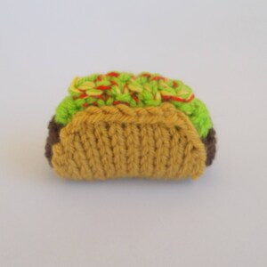 K&ouml;nnte beinhalten: Ein gestrickter Taco aus braunem, gelbem, gr&uuml;nem und rotem Garn. Der Taco ist mit gr&uuml;nem und rotem Garn gef&uuml;llt, das Salat und Tomate darstellt.