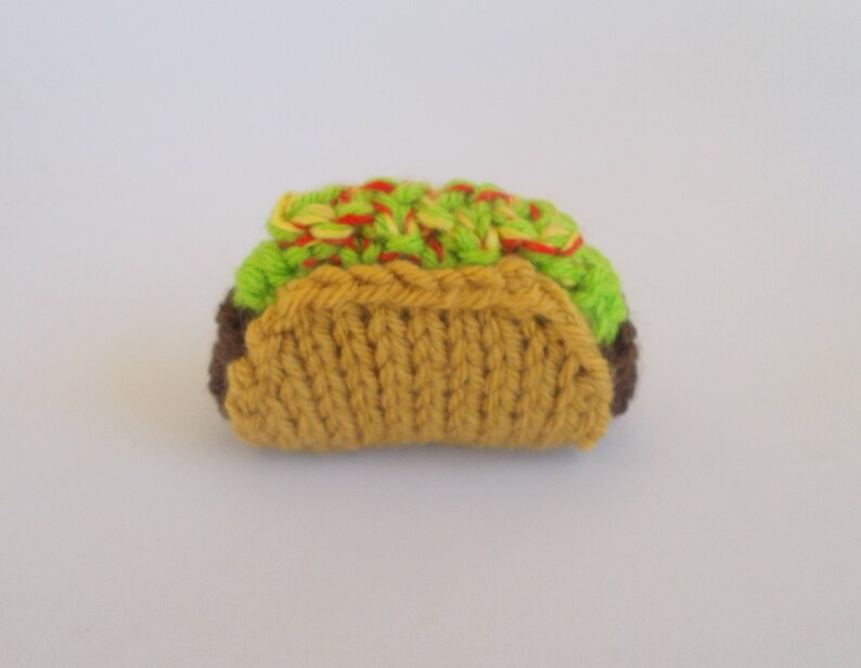 K&ouml;nnte beinhalten: Ein gestrickter Taco aus braunem, gelbem, gr&uuml;nem und rotem Garn. Der Taco ist mit gr&uuml;nem und rotem Garn gef&uuml;llt, das Salat und Tomate darstellt.