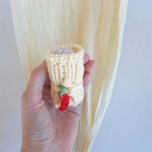 K&ouml;nnte beinhalten: Ein kleiner gestrickter Maiskolben mit einer roten Chilischote und gr&uuml;nen Bl&auml;ttern. Der Maiskolben ist gelb und die Chilischote ist rot. Der Maiskolben wird in einer Hand gehalten.