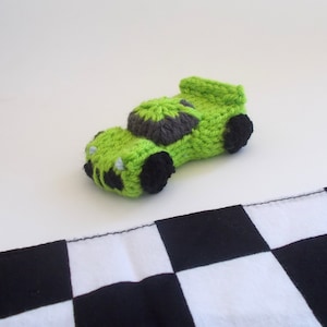 May include: A green knitted toy race car with black wheels on a black and white checkered surface.