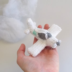 May include: A white knitted airplane toy with gray and green details. The airplane is held in a hand against a white background.