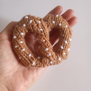 May include: A handmade, knitted pretzel in a warm brown color with white dotted accents. The soft, textured pretzel is held in a hand, showcasing the detailed craftsmanship of the item. A unique, food-themed item.