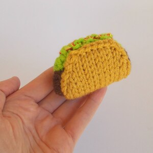 K&ouml;nnte beinhalten: Ein gestrickter Taco mit einer braunen Schale, gelber K&auml;sef&uuml;llung und gr&uuml;nem Salatbelag.