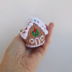 May include: A small knitted gingerbread house ornament with white frosting and colorful sprinkles. The house has a green wreath on the door and a brown door with white trim.