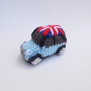 May include: A blue knitted toy car with a Union Jack flag on top. The car has black wheels and a gray knitted roof.