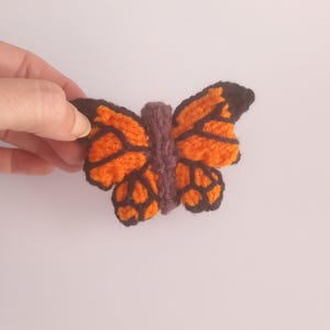 May include: A handmade, orange and black butterfly-shaped applique. The butterfly has a brown body and black outlines on the wings. The wings are orange with black details. The applique is held by a hand.