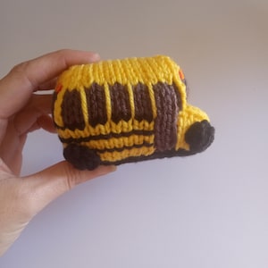 May include: A handmade, knitted yellow school bus toy with brown windows and black wheels. The bus has a yellow roof and is held in a person's hand. The bus is a small, soft toy.