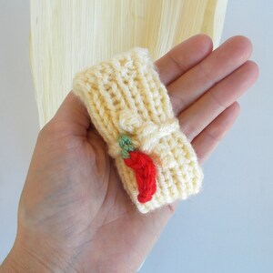 K&ouml;nnte beinhalten: Eine gestrickte gelbe Fingerpuppe mit einem roten Chili-Pfeffer-Design. Die Puppe wird in einer Hand gehalten.