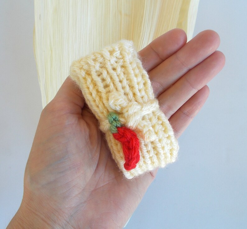 K&ouml;nnte beinhalten: Eine gestrickte gelbe Fingerpuppe mit einem roten Chili-Pfeffer-Design. Die Puppe wird in einer Hand gehalten.