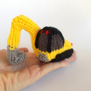 May include: A yellow knitted toy excavator with a gray arm and black cab. The excavator has red details on the cab.