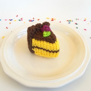 May include: A crocheted cake slice with brown frosting, yellow cake layers, and purple and green sprinkles on a white plate.