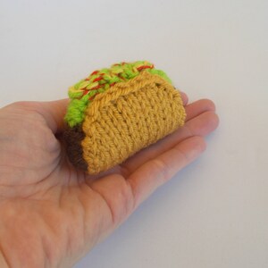 K&ouml;nnte beinhalten: Eine Hand h&auml;lt einen gestrickten Taco aus gr&uuml;nem, rotem und gelbem Garn. Der Taco ist so gestaltet, dass er einem echten Taco mit einer braunen F&uuml;llung &auml;hnelt.