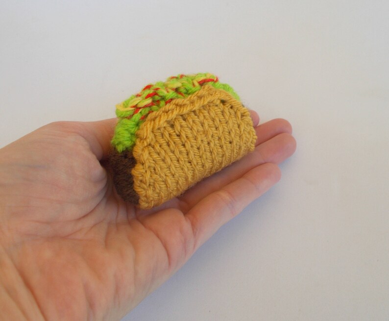 K&ouml;nnte beinhalten: Eine Hand h&auml;lt einen gestrickten Taco aus gr&uuml;nem, rotem und gelbem Garn. Der Taco ist so gestaltet, dass er einem echten Taco mit einer braunen F&uuml;llung &auml;hnelt.