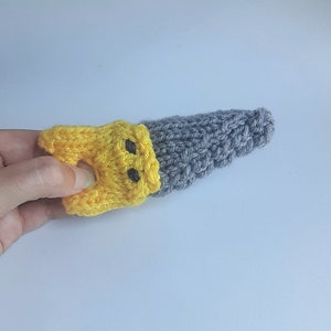May include: A yellow and gray knitted finger puppet with two black eyes. The puppet is shaped like a banana.