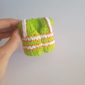 May include: A small, handmade, lime green crochet vest with white and orange stripes. The vest has white straps and orange trim. The item is held in a hand against a plain white background.