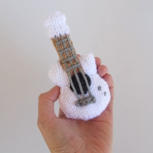 May include: A white knitted toy guitar with a brown and gray neck and black strings. The guitar is small enough to fit in the palm of a hand.