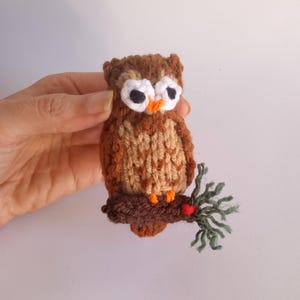 May include: A brown and tan knitted owl with white eyes and an orange beak perched on a brown branch with green leaves.