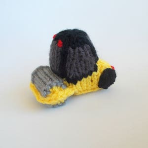 May include: A knitted toy bulldozer with a yellow body, gray tracks, and a black cab with red accents.