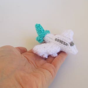 May include: A white knitted airplane toy with a teal tail fin. The airplane is small enough to fit in the palm of a hand.