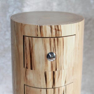 May include: A wooden cylindrical box with three drawers. The drawers have silver knobs and the wood has a natural, light brown color with dark brown streaks.