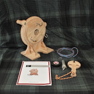 May include: A wooden rope making machine with a spool, a rope, a metal hook, a wooden handle, and a roll of tape. The machine is on a green and black plaid surface. The text on the paper says "Rope Making Machine Instructions".