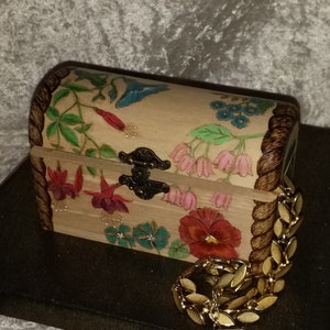 May include: A wooden box with a hinged lid, decorated with a floral design painted in bright colors, including pink, red, blue, and green. The box has a metal latch and is sitting on a black surface.