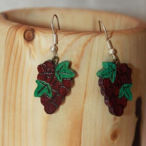 May include: A pair of wooden grape earrings. Each earring features a cluster of dark red grapes with green leaves. The earrings hang from silver-colored hooks and are displayed against a light-colored wooden background.