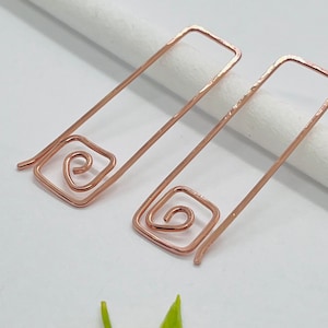 May include: A pair of rose gold earrings with a simple rectangular shape and a spiral design at the bottom.