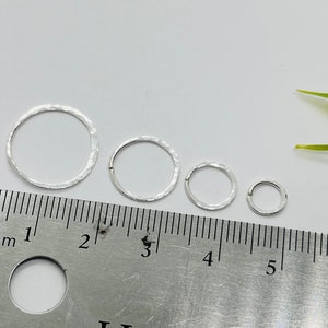 Puede incluir: Cuatro aros de plata martillados de diferentes tamaños, medidos en milímetros en una regla. Los aros tienen un diámetro de 1, 2, 3 y 4 centímetros.