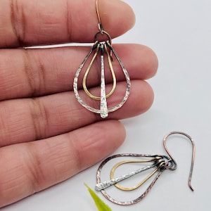 May include: Handmade earrings with a layered teardrop design. The earrings feature three nested teardrop shapes in silver, gold, and copper tones. The silver teardrop has a hammered texture. The earrings have a hook closure.