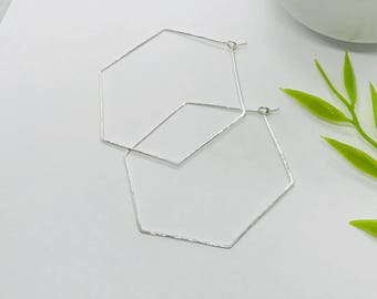 May include: A pair of geometric silver hoop earrings. Each earring is a six-sided polygon shape. The earrings are made of thin, textured metal and have a simple, minimalist design. The earrings are displayed on a white surface.