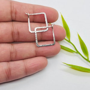 May include: A pair of silver square hoop earrings with a hammered texture. The earrings are displayed against a neutral background with a sprig of green leaves.
