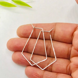 May include: A pair of silver geometric hoop earrings with a hammered texture. The earrings are in the shape of a diamond and are shown in a hand.