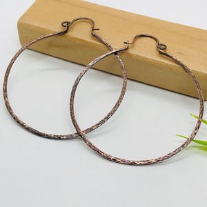 May include: A pair of large, hammered copper hoop earrings. The earrings have a textured surface and a warm, reddish-brown color. They are displayed on a light brown wooden block against a white background, with a touch of green foliage.